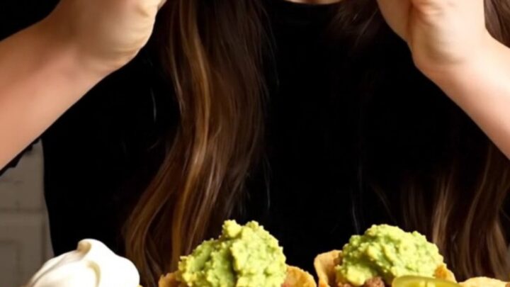 Golden baked mini taco cups topped with sour cream, guacamole and pickled jalapeño slices, one lifted with a long cheese pull
