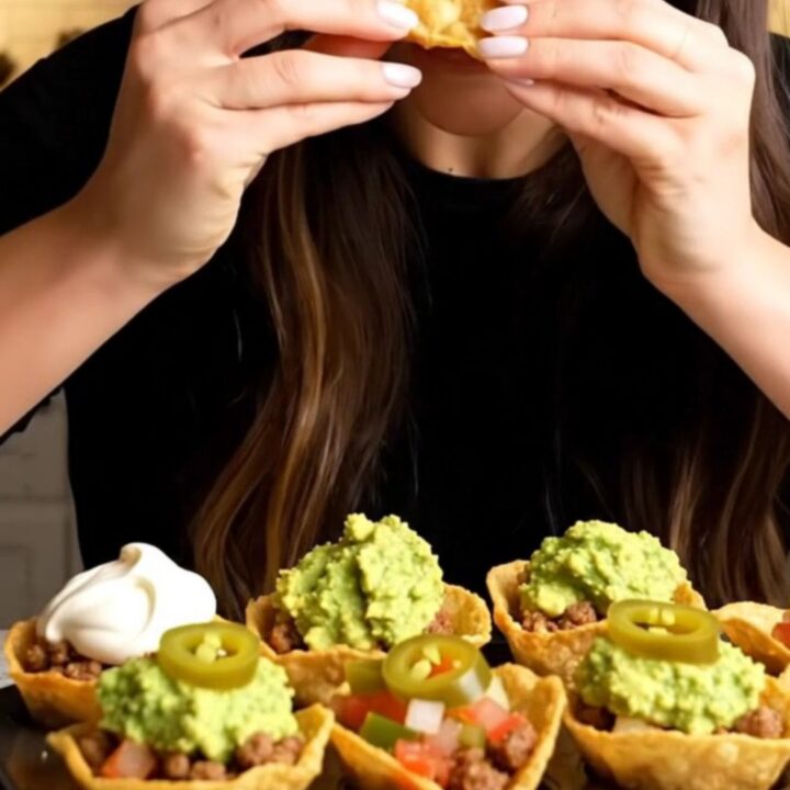 Golden baked mini taco cups topped with sour cream, guacamole and pickled jalapeño slices, one lifted with a long cheese pull