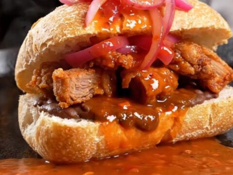 Torta ahogada in a shallow bowl, drowned in bright red chile de árbol tomato sauce and topped with lime-pickled red onions
