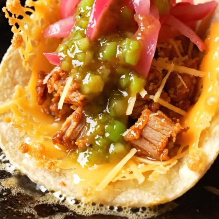 Crispy carnitas taco wrapped in a golden lacy cheese skirt, snapped in half to show crackling edges and juicy pork