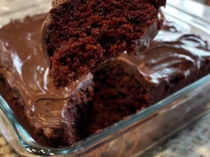 A moist slice of chocolate wacky cake with glossy frosting lifted from a glass baking dish