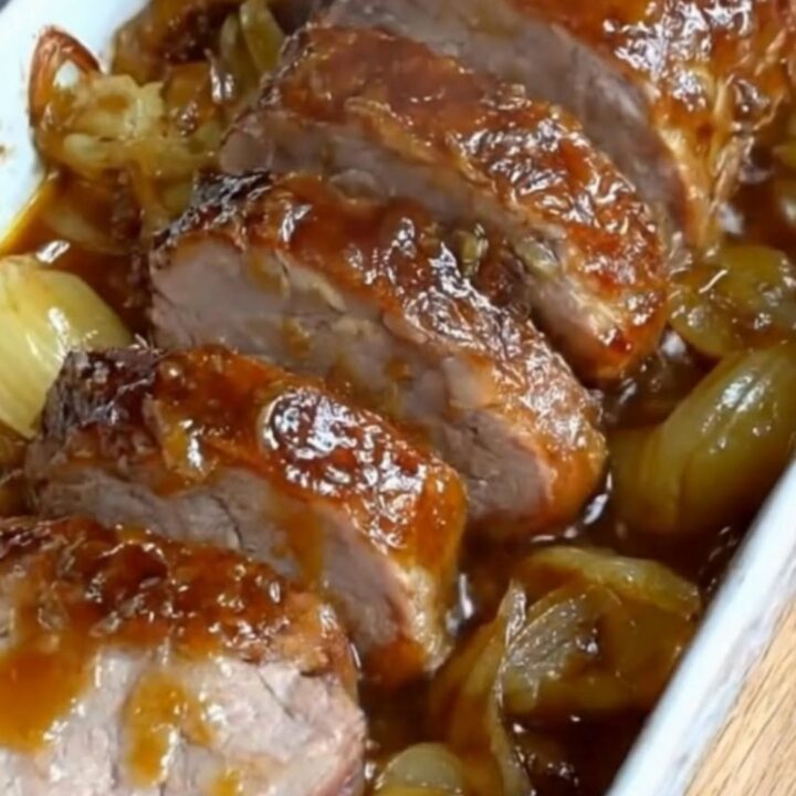 Sliced glazed pork tenderloin covered in caramelized onions lifted from a white ceramic baking dish with tongs