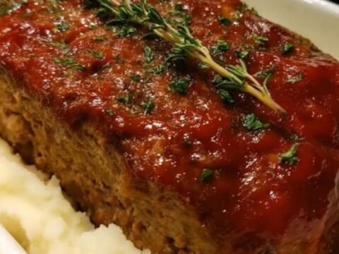A glazed meatloaf with glossy red glaze nestled in creamy mashed potatoes in a white baking dish, steam rising