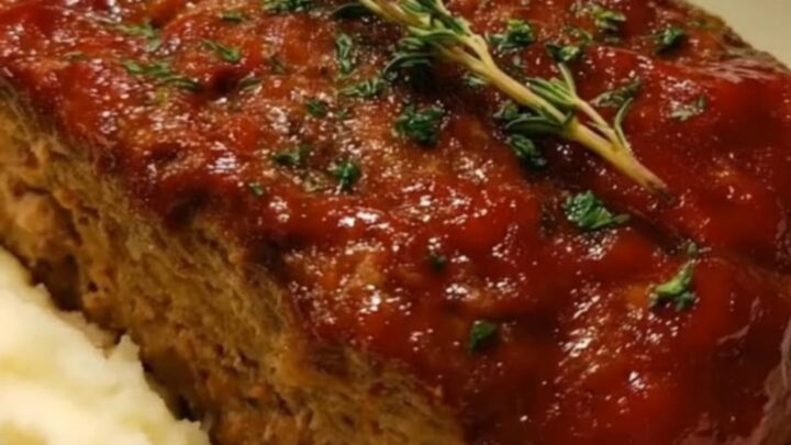 A glazed meatloaf with glossy red glaze nestled in creamy mashed potatoes in a white baking dish, steam rising