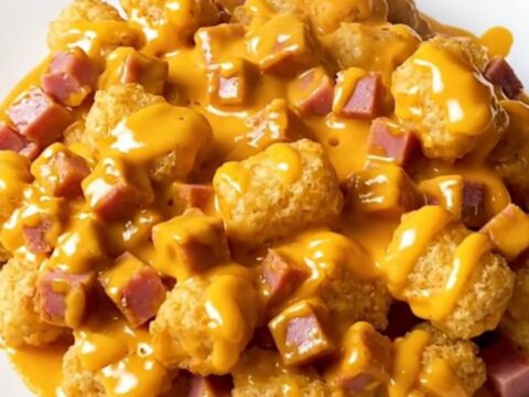 Slow cooker ham and potato tot casserole with melted cheddar sauce in a cream stoneware crock on a beige countertop