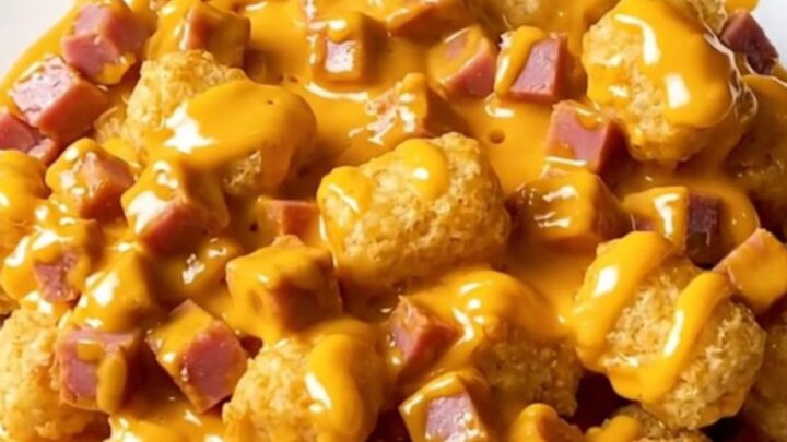 Slow cooker ham and potato tot casserole with melted cheddar sauce in a cream stoneware crock on a beige countertop