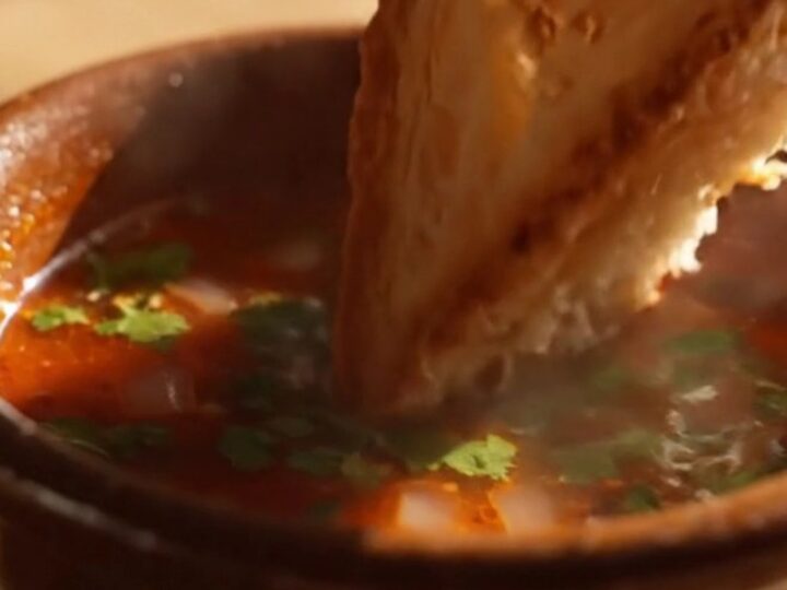 Birria grilled cheese pulled apart in slow motion, golden cheese strings soaked in glossy red birria oil between thick