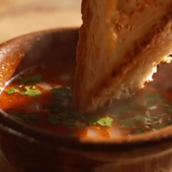 Birria grilled cheese pulled apart in slow motion, golden cheese strings soaked in glossy red birria oil between thick
