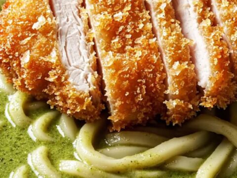 Sliced crispy panko chicken katsu over thick udon noodles in glossy emerald green curry coconut broth, topped with Thai basil