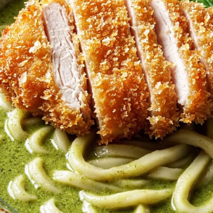 Sliced crispy panko chicken katsu over thick udon noodles in glossy emerald green curry coconut broth, topped with Thai basil