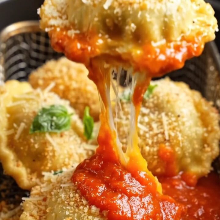 Golden toasted air fryer ravioli piled on a platter with grated Parmesan, fresh basil and a bowl of warm marinara for dunking