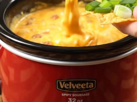 Glossy slow cooker sausage queso stretching in a thick ribbon from a tortilla chip over a crimson ceramic crock