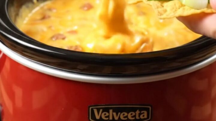 Glossy slow cooker sausage queso stretching in a thick ribbon from a tortilla chip over a crimson ceramic crock