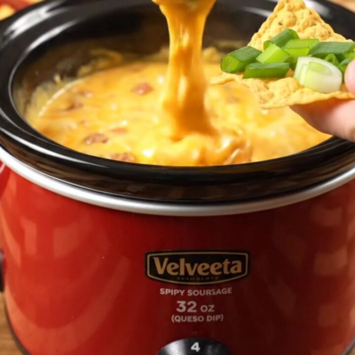 Glossy slow cooker sausage queso stretching in a thick ribbon from a tortilla chip over a crimson ceramic crock