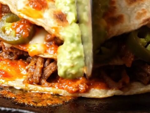 Double-stacked lamb birria pizzadilla cut into wedges with oozing Oaxaca cheese and avocado salsa drizzle