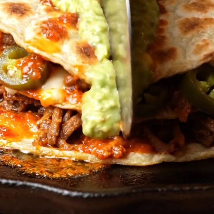 Double-stacked lamb birria pizzadilla cut into wedges with oozing Oaxaca cheese and avocado salsa drizzle