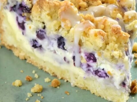 Gâteau crumble aux myrtilles coupé en carré sur une assiette vert sauge, couche cheesecake visible et streusel doré