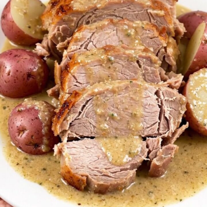 Sliced creamy ranch pork roast with halved red potatoes and glossy sauce on a white plate, steaming
