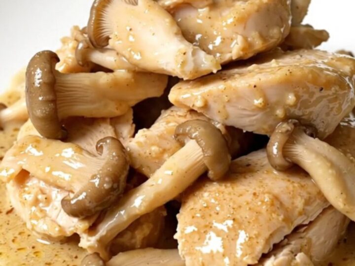 Tender slow cooker chicken breasts on a white plate under a glossy cream and oyster mushroom sauce, steaming