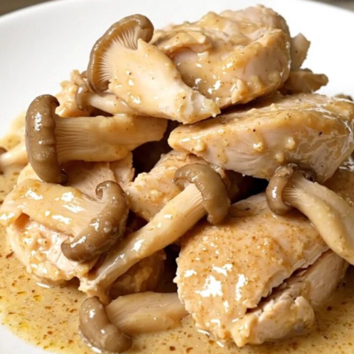 Tender slow cooker chicken breasts on a white plate under a glossy cream and oyster mushroom sauce, steaming