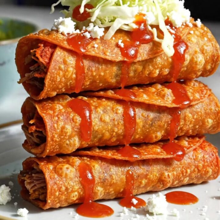 Stacked crispy birria taquitos topped with iceberg, queso fresco and consomé drizzle beside a bowl of green avocado salsa