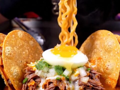 Steaming birria ramen in a black ceramic bowl with a jammy egg, cilantro, and two crispy quesatacos propped on the rim