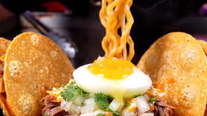 Steaming birria ramen in a black ceramic bowl with a jammy egg, cilantro, and two crispy quesatacos propped on the rim