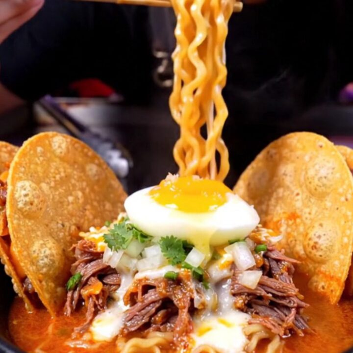 Steaming birria ramen in a black ceramic bowl with a jammy egg, cilantro, and two crispy quesatacos propped on the rim