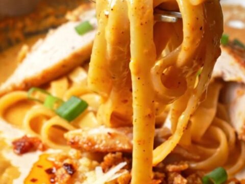 A fork twirling fettuccine coated in creamy chili crisp Alfredo sauce, topped with sliced chicken and scallions.