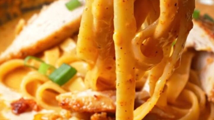 A fork twirling fettuccine coated in creamy chili crisp Alfredo sauce, topped with sliced chicken and scallions.