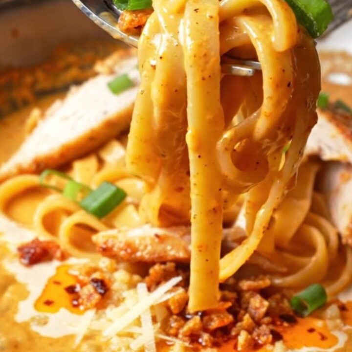 A fork twirling fettuccine coated in creamy chili crisp Alfredo sauce, topped with sliced chicken and scallions.
