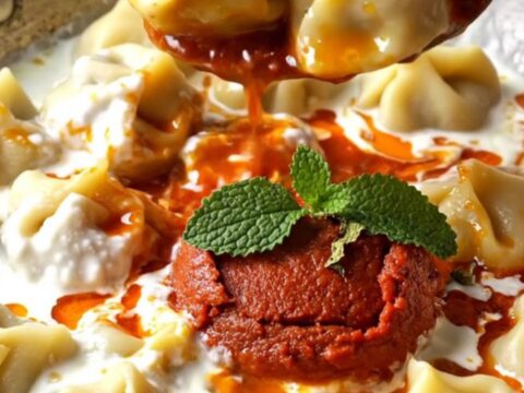 A bowl of Turkish mantı dumplings topped with garlic yogurt and red paprika butter, garnished with dried mint.