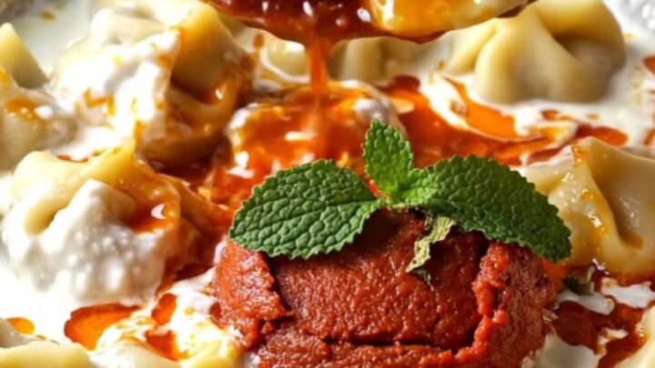 A bowl of Turkish mantı dumplings topped with garlic yogurt and red paprika butter, garnished with dried mint.