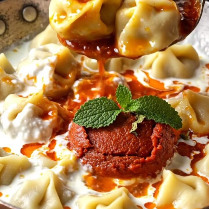A bowl of Turkish mantı dumplings topped with garlic yogurt and red paprika butter, garnished with dried mint.