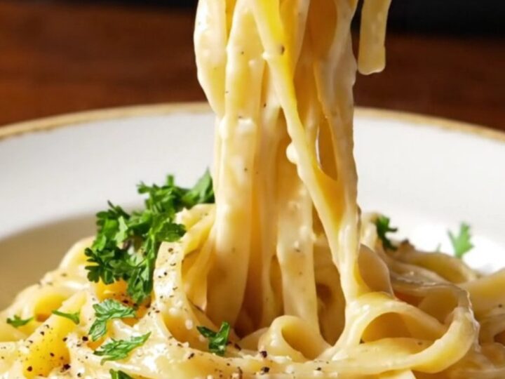 A fork twirling glossy fettuccine Alfredo with creamy sauce and steam rising in a shallow bowl.
