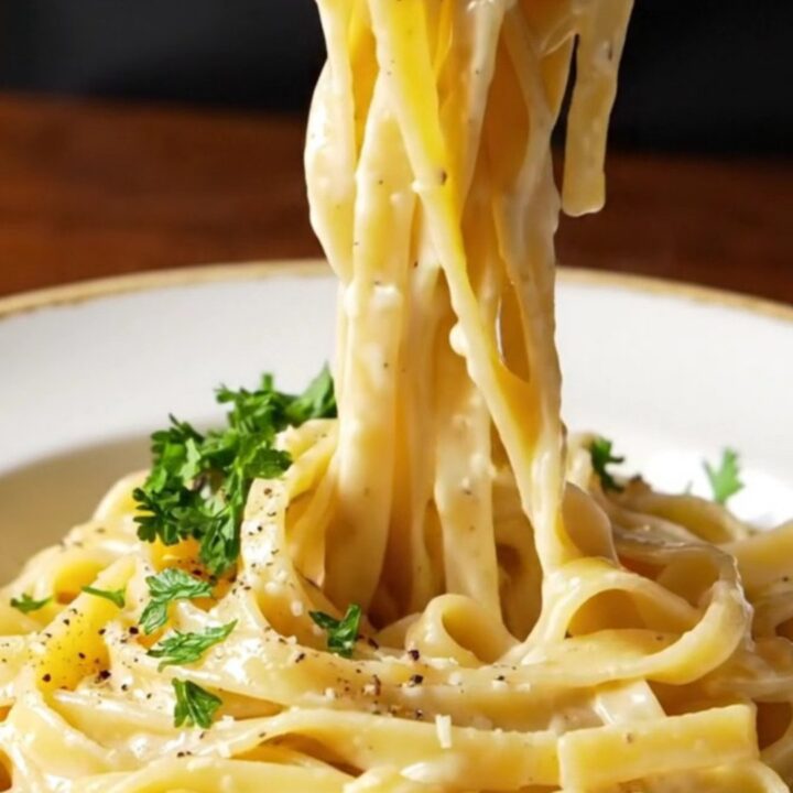 A fork twirling glossy fettuccine Alfredo with creamy sauce and steam rising in a shallow bowl.