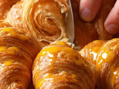 Golden flaky croissants stacked on a cooling rack, with visible layers and a glossy crust.