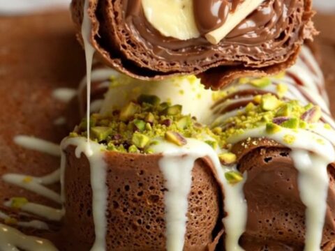 Sliced dessert crepe sushi rolls topped with white chocolate drizzle and pistachios on a platter