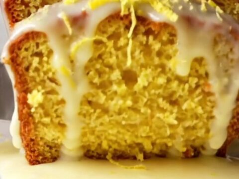 A thick slice of glazed lemon loaf cake on a plate, showing the moist yellow crumb and white sugar glaze with lemon zest