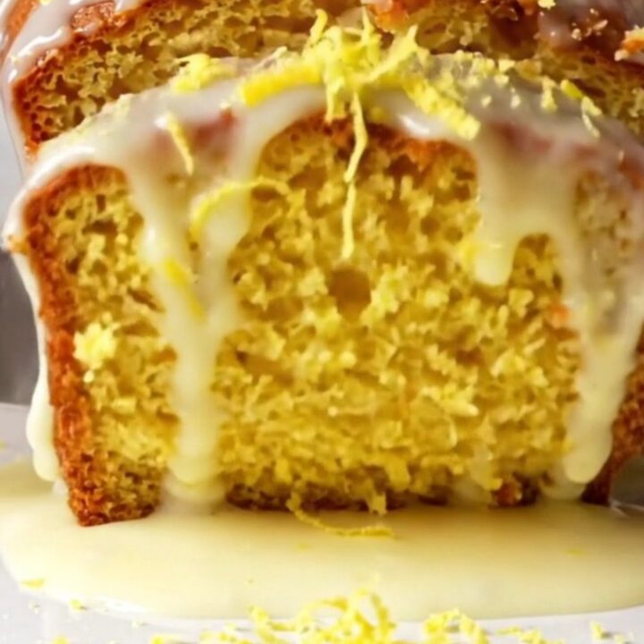 A thick slice of glazed lemon loaf cake on a plate, showing the moist yellow crumb and white sugar glaze with lemon zest
