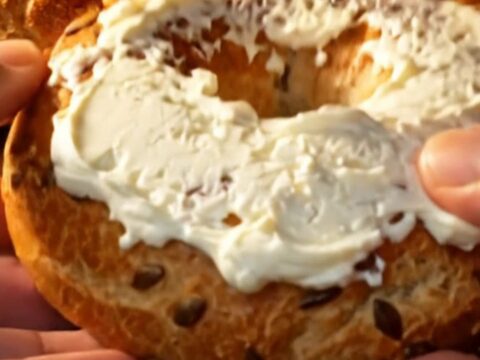 Hands tearing open a golden seed-crusted bagel, steam rising from the chewy crumb spread with cream cheese