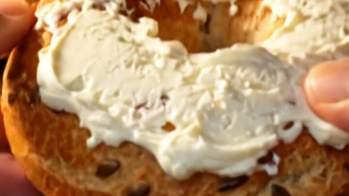 Hands tearing open a golden seed-crusted bagel, steam rising from the chewy crumb spread with cream cheese