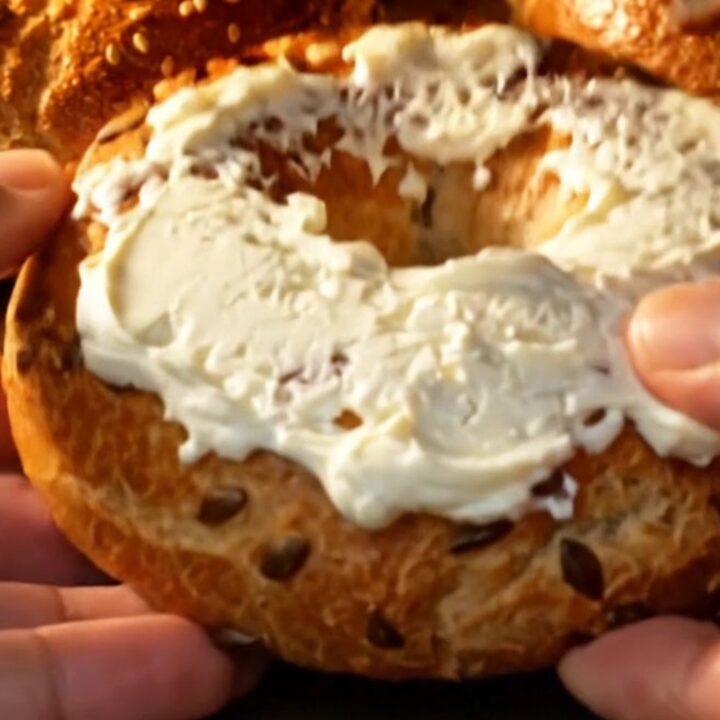 Hands tearing open a golden seed-crusted bagel, steam rising from the chewy crumb spread with cream cheese
