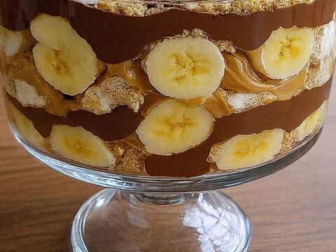 A stunning chocolate peanut butter banana trifle in a footed glass bowl, topped with whipped cream and peanuts.