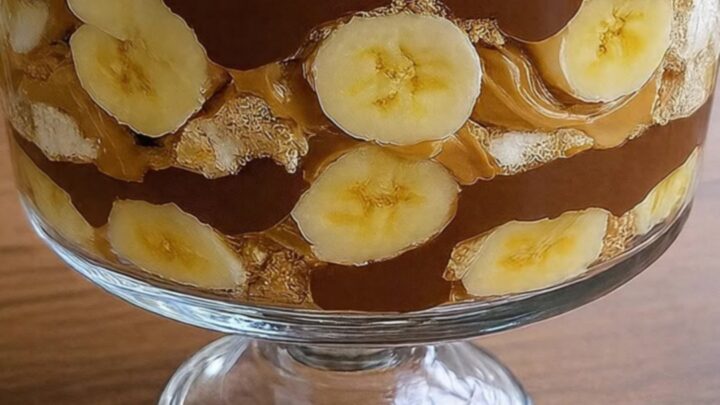 A stunning chocolate peanut butter banana trifle in a footed glass bowl, topped with whipped cream and peanuts.