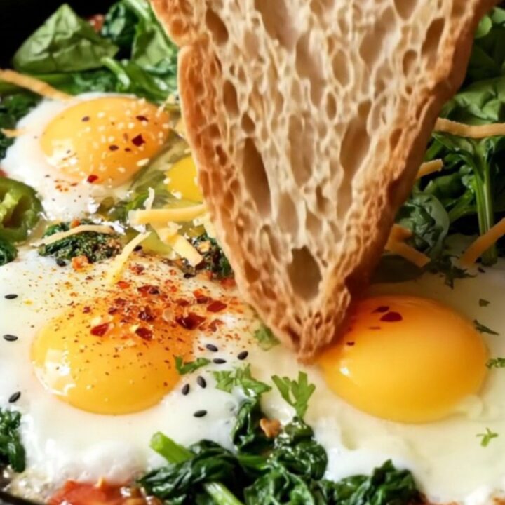 Cast-iron skillet with baby spinach, blistered tomatoes, eggs, and melted cheese, garnished with parsley and nigella seeds
