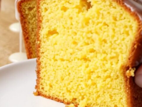 Golden whole lemon loaf cake with white glaze on a cutting board, slice cut showing moist yellow crumb.