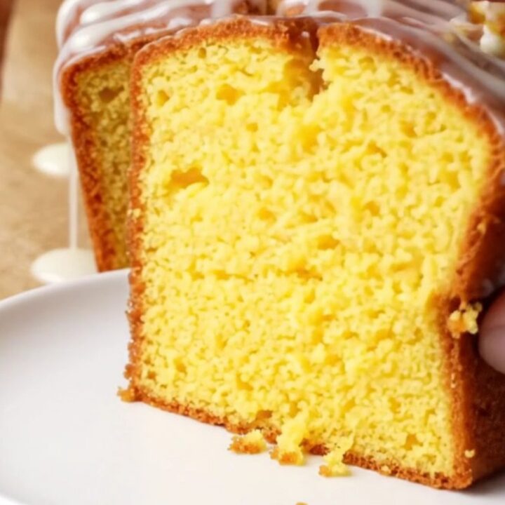 Golden whole lemon loaf cake with white glaze on a cutting board, slice cut showing moist yellow crumb.