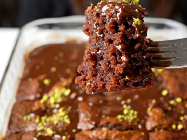 A square of chocolate cake soaked in glossy fudge sauce, topped with crushed pistachios, on a plate.