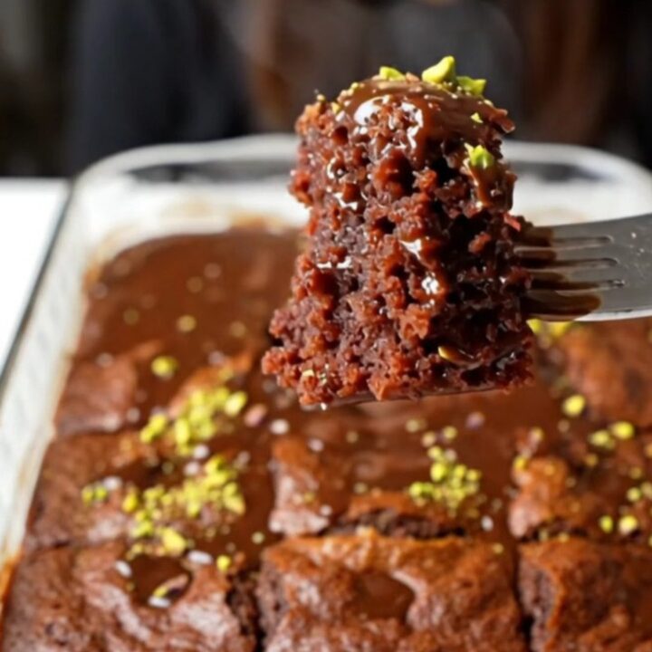 A square of chocolate cake soaked in glossy fudge sauce, topped with crushed pistachios, on a plate.